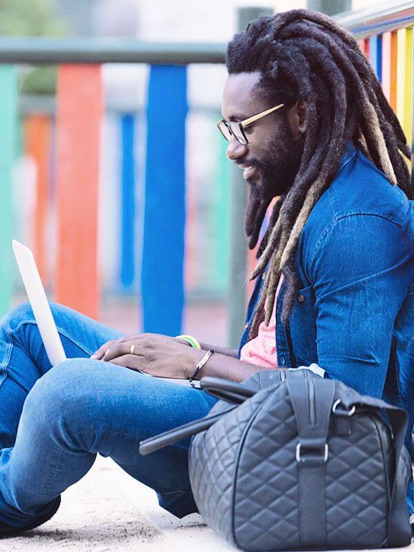 A man wearing denim sits on the ground by a multi-colored fence, using his laptop.